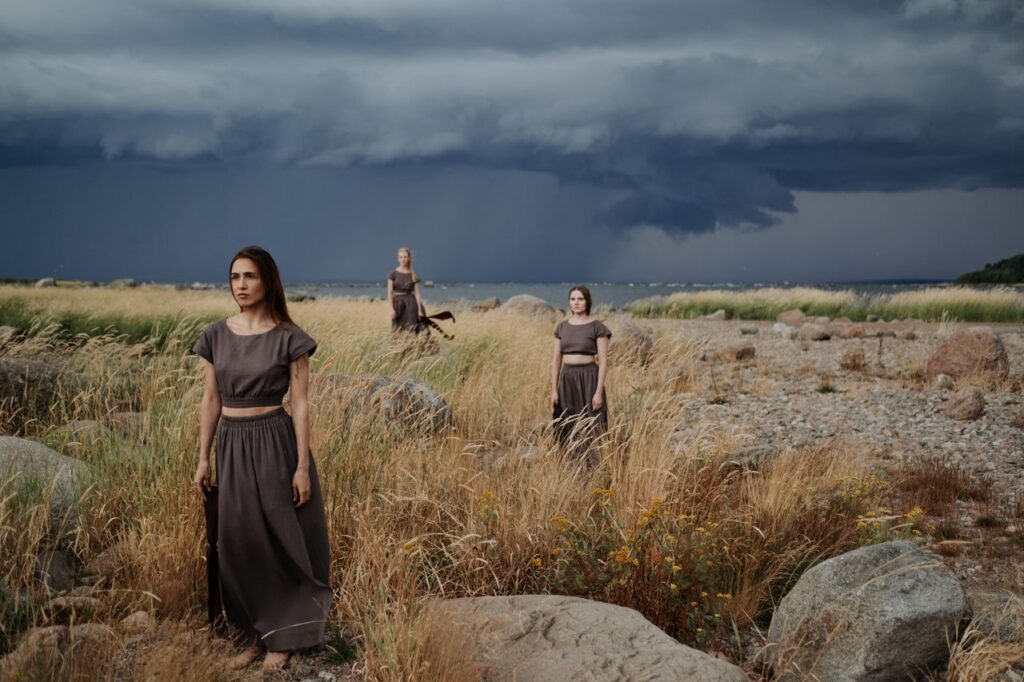 Three women in long dresses standing in a rocky grass field under a stormy sky.