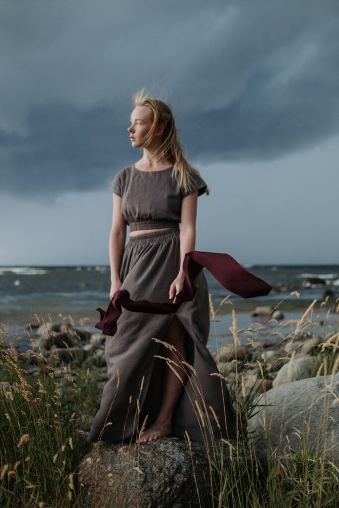 Woman standing on rocky shore with stormy sky, wearing a long flowing dress.