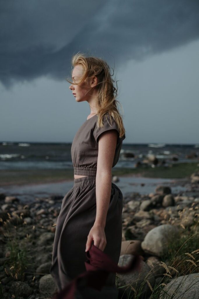 A woman stands by a rocky seashore under a moody, stormy sky, embodying solitude and introspection.