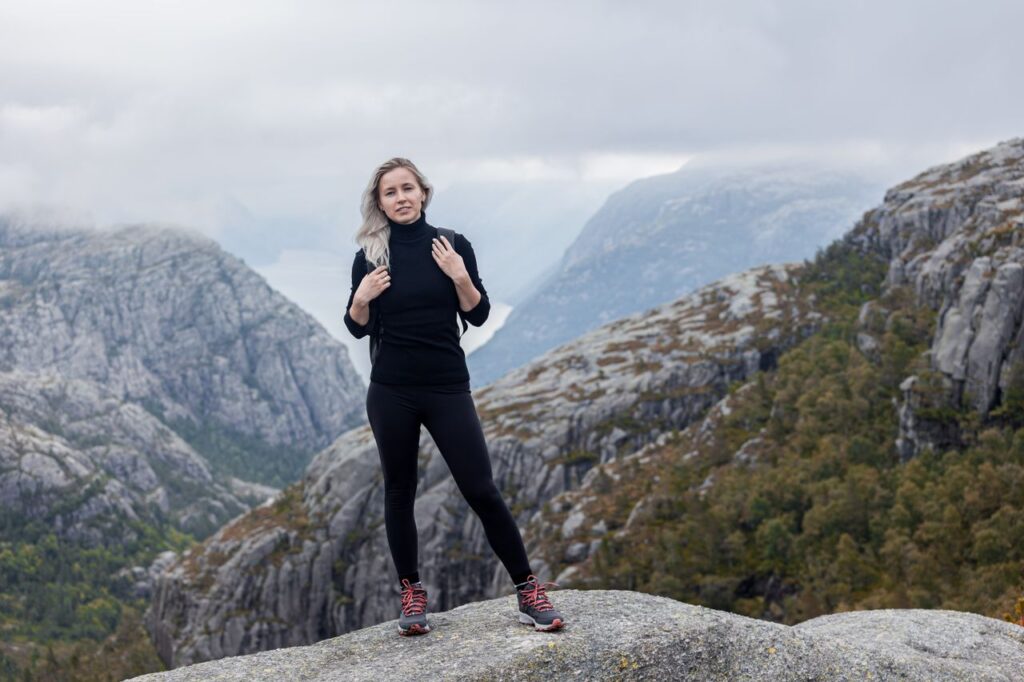Blonde woman in black clothing standing confidently on a rocky mountain landscape.
