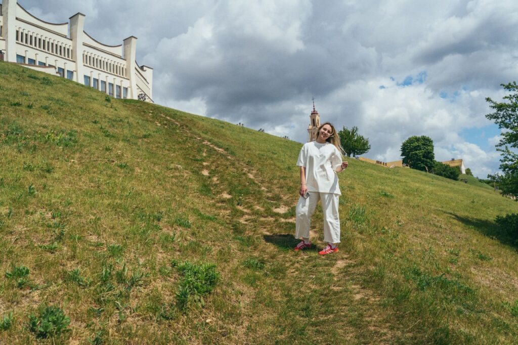 A person stands on a grassy hill on a sunny day with a cloudy sky, near historic architecture.