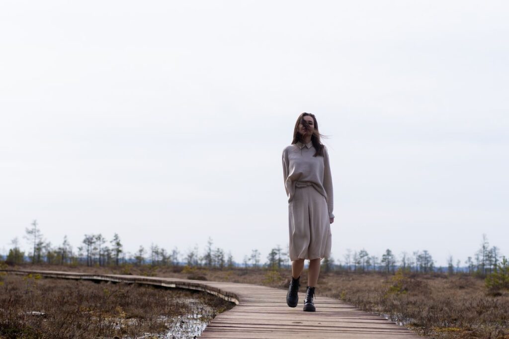 Full body of young female with flying hair wandering on timber path surrounded with dry grass under cloudy sky
