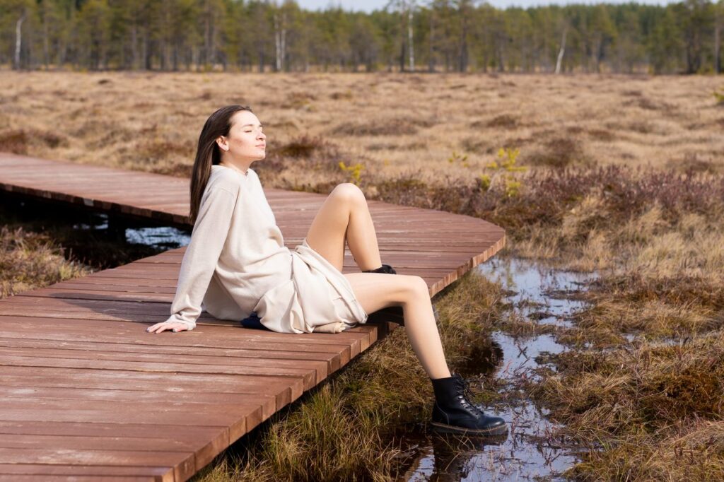 Full body of positive young female with eyes closed smiling on timber path near pools with grass in spring
