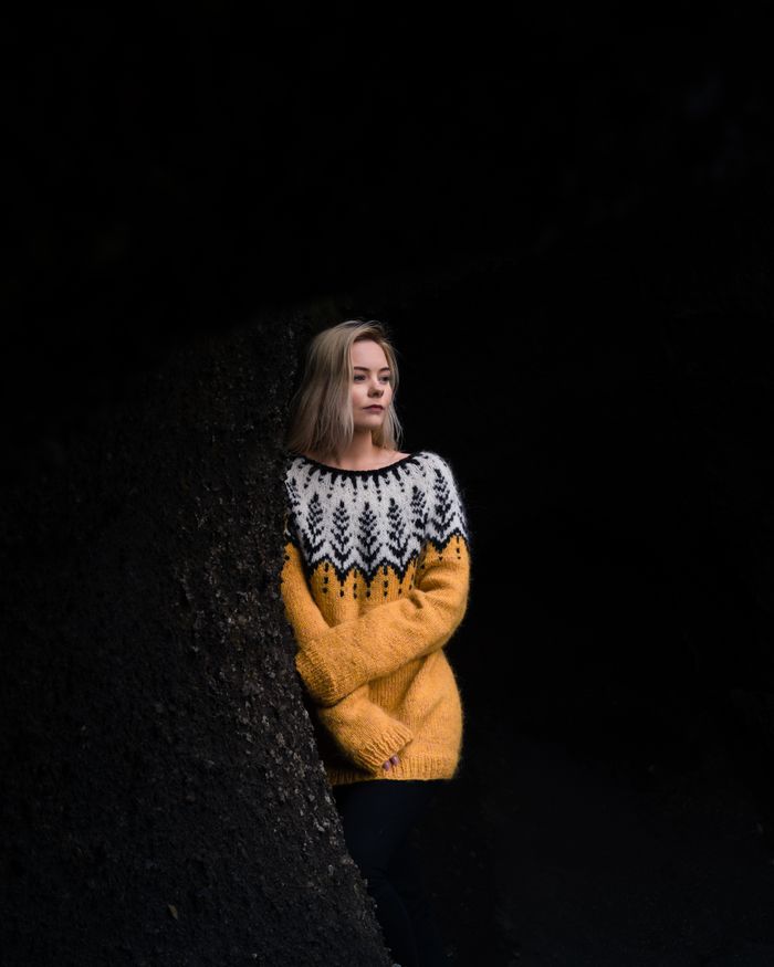 Woman in a yellow Icelandic sweater standing against a dark, textured backdrop, reflecting solitude.